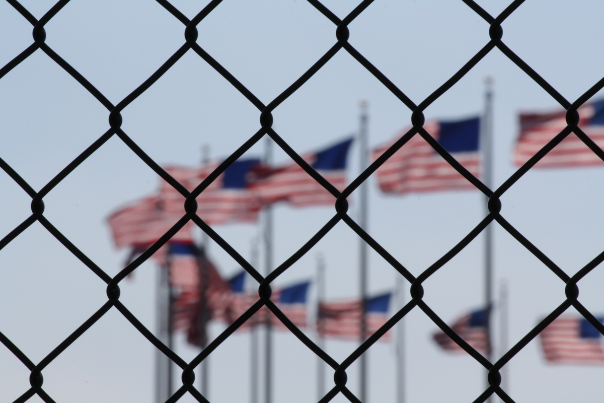 A close-up image of a chain-linked fence with American flags in the background.